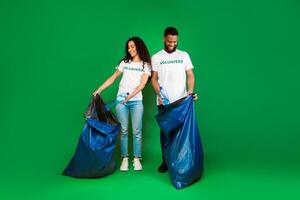 Two Multiracial Volunteers With Big Blue Garbage Bags Picking Used Plastic Litter And Trash Standing Over Green Studio Background. Ecology, Volunteering Work, Keep Environment Clean. Full Length photo