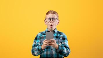 Surprised schoolboy playing game or using amazing application on smartphone over yellow studio background, copy space. Amazed ginger kid using newest mobile phone, entertainment concept photo