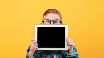 Amazed kid in eyeglasses hiding behind digital tablet with empty screen over yellow studio background. Excited redhead schoolboy showing pad, mockup, advertisement for cute application photo