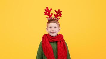 Smiling kid in green winter sweater and red scarf posing on yellow studio background, copy space. Happy ginger boy with deer accessory on his head waiting for Christmas and New Year photo