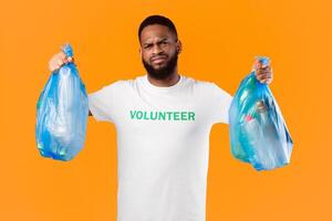 Discontented Black Volunteer Man Holding Two Plastic Garbage Bags Full Of Litter And Bottles Standing Over Yellow Studio Background. Waste Sorting, Junk Ecology Problem, Volunteering Work Concept photo
