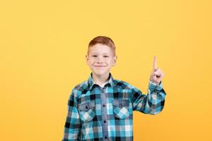 Handsome redhead boy in casual outfit pointing up at empty space for text or advertising over yellow studio background. Smiling kid showing exciting advertisement, aiming at free space photo