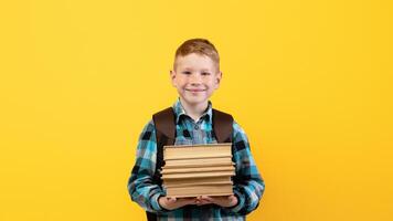 Smiling schoolboy with backpack holding heap of books, yellow studio background, copy space. Happy boy going to school, holding books, enjoying his school days. Kids education concept photo