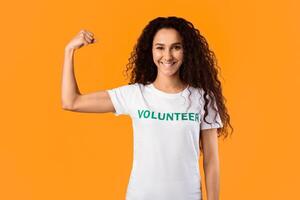Power Of Kindness. Volunteer Lady Posing Showing Biceps Muscles Standing Over Yellow Studio Background, Smiling To Camera. Volunteering Missions, Charity And Voluntary Job Concept photo