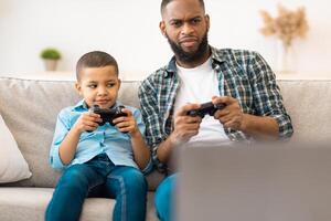 Young Black Father And Little Son Playing Games Sitting With Gamepad Controllers At Home. Family Bonding Having Fun, Dad And Kid Play Videogames Together On Weekend photo