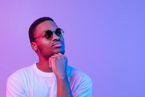 Portrait Of Pensive African American Guy In Stylish Eyeglasses Looking Away, Young Black Man Thinking About Something While Posing Under Neon Light Over Purple Background, Panorama With Free Space photo