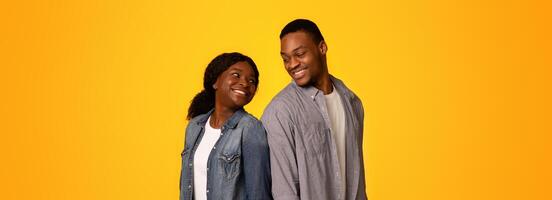 Cheerful black man and woman standing back to back and turning heads to each other while standing isolated over yellow background, young african american lovers smiling and having fun, copy space photo