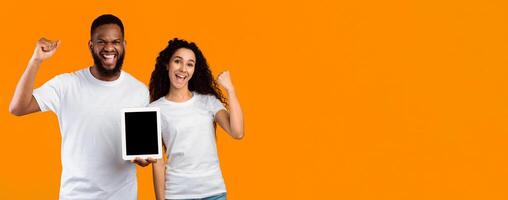 Diverse Couple Showing Tablet Empty Screen Shaking Fists Celebrating Good News Standing Over Yellow Studio Background, Smiling To Camera. Great Mobile Application. Mockup, Panorama With Blank Space photo