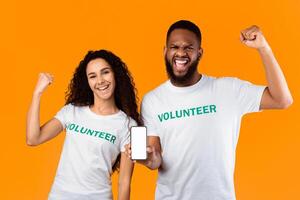 Volunteering. Two Diverse Volunteers Showing Smartphone With Blank Screen Recommending Volunteering App Standing On Yellow Studio Background. Power Of Kindness Concept. Mockup photo
