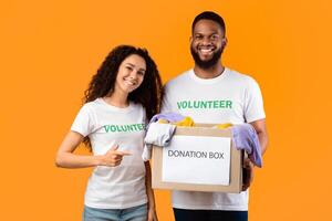 Two Cheerful Volunteers Holding Donation Box For Charity Standing Posing On Yellow Studio Background, Pointing Finger At Container With Clothes For Homeless People. Volunteering Work Concept photo