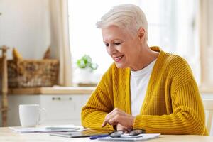 Positive grey-haired senior woman using digital tablet in kitchen, taking notes, counting month spending, copy space. Smiling old lady grandmother enjoying nice mobile application photo