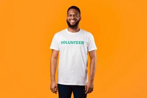 Happy Black Volunteer Guy Standing Smiling To Camera Wearing Uniform T-Shirt With Text On Yellow Background. Activist Posing In Studio. Volunteering Career Concept photo