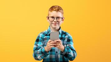 Smiling boy schooler playing game or using amazing application on smartphone over yellow studio background, copy space. Happy ginger kid in glasses using newest mobile phone, entertainment concept photo