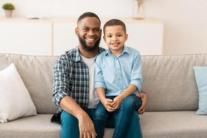 Portrait Of Happy Black Father And Son, Little Boy Sitting On Dad's Knees Smiling To Camera At Home. Cheerful Young Daddy And His Kid Posing Indoor. Family, Happiness And Childhood Concept photo