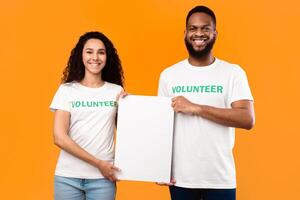 Two Diverse Volunteers Holding Empty Paper Poster For Your Text, Smiling To Camera Standing Over Yellow Studio Background. Volunteering Work, Charity Advertisement Banner. Mockup photo