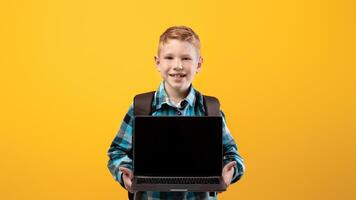 Cheerful ginger kid with schoolbag showing laptop with blank screen on yellow studio background, copy space. Happy schoolboy holding modern laptop, using technologies in education, mockup photo