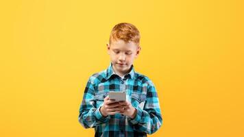 Cute ginger kid with mobile phone on yellow studio background, using exciting mobile applications or chatting with friends, copy space. Happy redhead boy using smartphone, gadget addiction photo