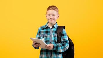 Technologies in school education. Smiling smart schoolboy with backpack holding digital tablet over yellow studio background, copy space. Cute kid using pad for studying, doing homework, websurfing photo