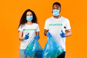 Two Multiethnic Volunteers Holding Plastic Bottles And Blue Garbage Bags With Litter Standing On Yellow Studio Background. Waste Sorting And Plastic Disposal. Keep Planet Clean Concept photo