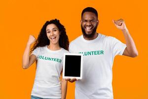 Two Multicultural Volunteers Showing Tablet With Blank Screen Gesturing Yes Standing Over Yellow Studio Background, Smiling To Camera. Activists Recommending Charity Application. Mockup photo