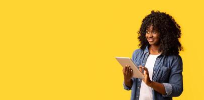 Joyful african american young woman with newest digital tablet looking at copy space on yellow studio background and smiling, panorama. Advertisement for mobile application concept photo