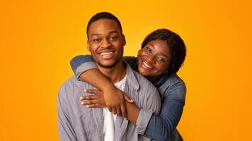 Portrait of young cheerful black couple hugging and smiling at camera while standing isolated over yellow background, loving african american woman embracing her boyfriend from back, copy space photo