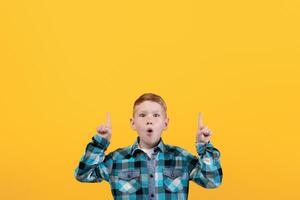 Shocked kid showing exciting advertisement, aiming at free space for text or advertisement. Surprised redhead schoolboy pointing up at empty space over yellow studio background photo