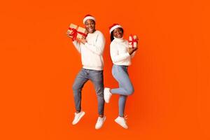 Cheerful Black Couple In Santa Hats Posing With Christmas Gifts, Holding Wrapped Present Boxes And Smiling At Camera While Jumping In Air Over Orange Studio Background, Full Length, Copy Space photo