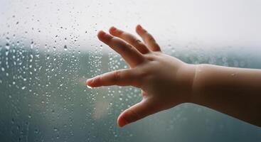 Small child hand reaching out to touch a single raindrop on a wet windowpane photo