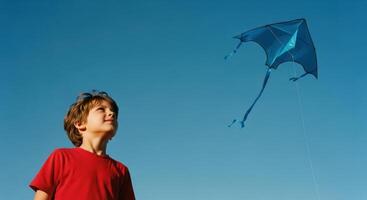 Young boy in red shirt looking up at a blue kite flying high in a clear sky photo