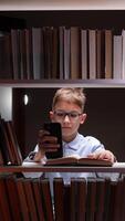 A boy in glasses is looking at a phone while sitting on a shelf of books video