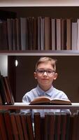 A boy in glasses is reading a book in front of a shelf of books video