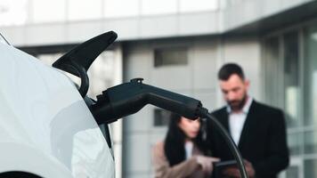 A business woman with a tablet in hand discusses project details with a business man while their electric car is charging in the parking lot. Concept of modern cars, business travel. video