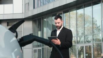 A confident businessman next to his electric car waiting for charging to finish. The concept of clean energy and sustainable development, emphasizing the importance of green technology video