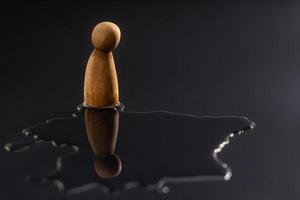 A wooden peg doll stands alone, staring down at its reflection in a puddle. Symbolizes symbolizing mental health crisis, deep depression, stress, and emotional pressure photo