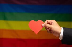 Hand holding a red heart shape symbol made from paper against a rainbow flag background photo