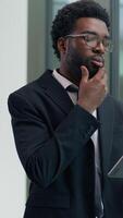 Vertical view African American pensive thinking business man in office looking gazing out window using digital tablet computer device smiling businessman guy creative worker find idea browsing screen video