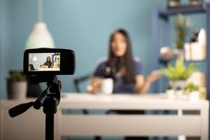 Asian woman blogger seated at desk in a professional studio setting, holding a cup of coffee and speaking into a microphone. The image emphasizes digital content creation and filming equipment. photo