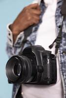 Closeup shot of a professional camera with its strap held by african american individual. Selective focus on a DSLR hanging over a white shirt and denim jacket, highlighting its fine details. photo