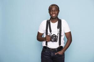 Smiling black photographer in suspenders standing confidently in a studio, posing with a camera around his neck. The clean blue background enhances the sharp, professional look. photo