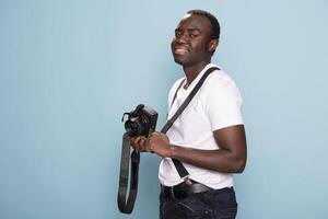 African american man stands confidently, holding a camera in one hand while adjusting his suspenders strap with the other. Portrait of black male model in casual attire, posing with his DSLR. photo
