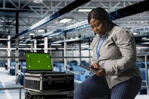 Mockup laptop next to female engineer in server hub using tablet to optimize hardware performance. African american woman in data center using green screen notebook, upgrading rackmounts photo
