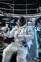 Cheerful female astronaut doing videocall with command center on Earth using smartphone. Happy woman in orbit station talks with ground control researchers using mobile phone photo