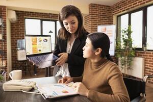 Caucasian employee holds a laptop and receiving strategic advice from her asian manager in brick walled office. Two young businesswomen collaborate using digital tools and documents. photo
