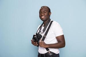 African american photographer posing with a relaxed yet confident expression, gripping his suspenders and holding his camera. The solid blue background adds a modern and clean look. photo