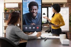 Female consultant stands with tablet, and coworker sits at desk, presenting company strategy ideas to male client during virtual meeting. Businesswomen engaged in videoconference call with black man. photo