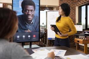 African american man on digital monitor, having financial discussion via call with businesswomen in startup office. Asian manager standing with tablet, presenting weekly reports to her coworkers photo