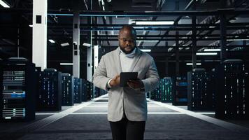 IT professional looking over data center checkup checklist paperwork on tablet. African american man walking in server farm, reading documentation files on device, camera A photo