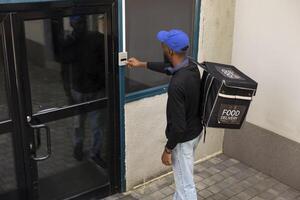 African American delivery man arrives at the customer doorstep with meal package, using the intercom and door bell to call on the client. Express delivery quick service, courier bag. photo