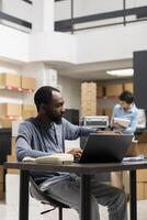 Black male executive manager uses laptop for order logistics in the storage room, working in a local distribution hub. Supply room filled with boxes and inventory goods, e commerce. photo